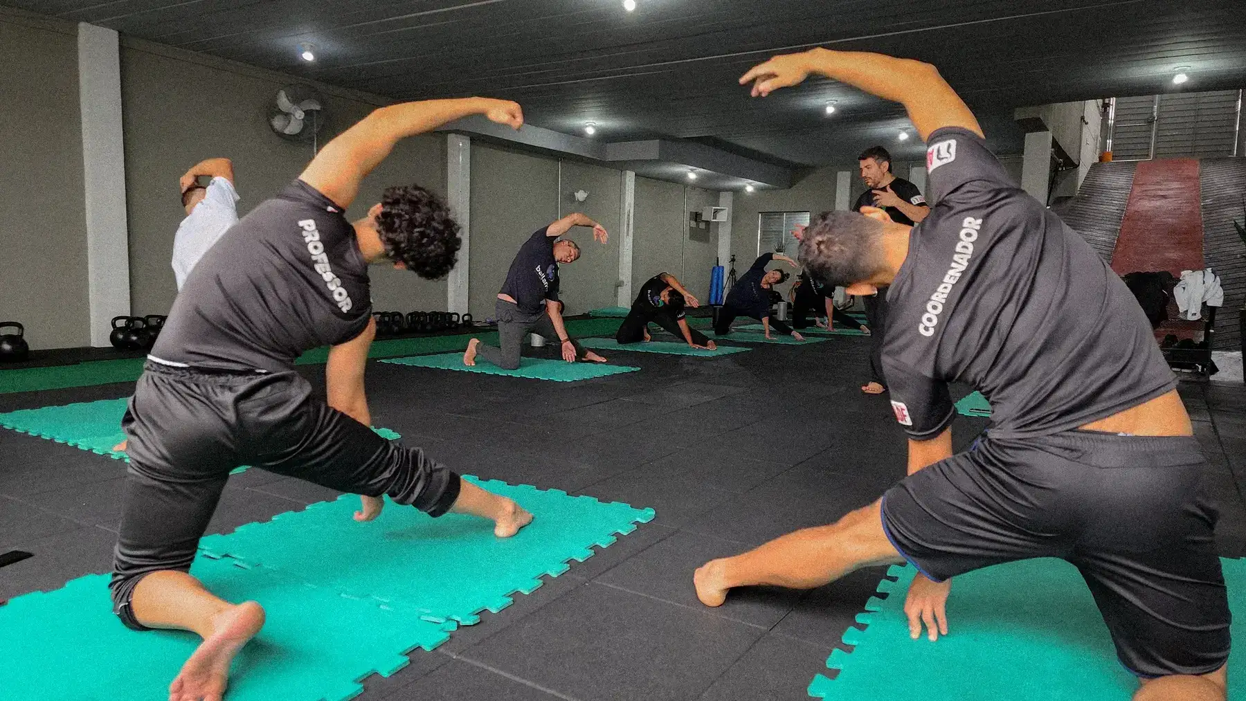 Professor e aluno em alongamento lateral, com a turma ao fundo